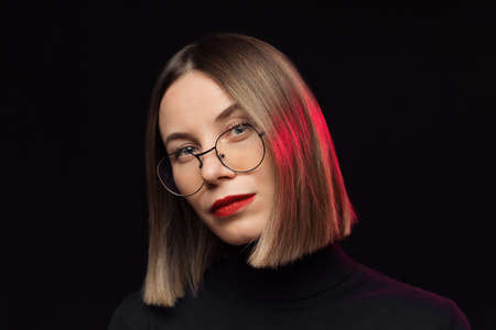 Close up woman portrait wearing glasses with black frames, with red lips, looking to camera on black...