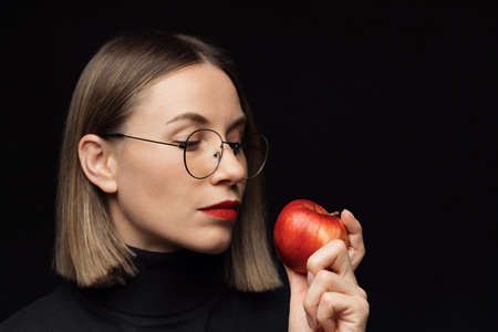 Close up woman portrait wearing glasses with black frames, with red lips, looking to a red apple on black background with focused face expression. Mocupの写真素材