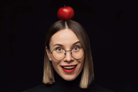 Close up surprised woman portrait wearing glasses with black frames, with red lips, looking at the camera with a red apple on the head over black background.の写真素材