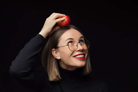 Close up surprised woman portrait wearing glasses with black frames, with red lips, looking at the camera with a red apple on the head over black background.の写真素材