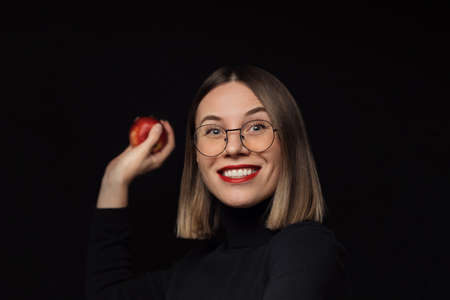 Excited woman wearing glasses with black frames, with red lips, looking at the camera wants to throw a red apple over black background.の写真素材