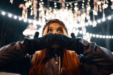 Outdoor night photo of young beautiful happy smiling girl enjoying festive decoration, posing in Christmas fair, in street of european city, wearing knitted beanie hatの写真素材