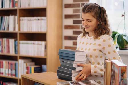 Teen girl among a pile of books. A young girl holding books with shelves in the background. She is...