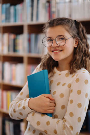 Teen girl among a pile of books. A young girl holding book with shelves in the background. She is...