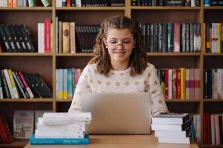 Attractive happy young girl student studying at the college library, sitting at the desk, using laptop computer, having video chatの写真素材