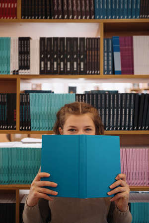 Portrait of a pretty young girl hiding behind an open blue book and looking away over bookshelf backgroundの写真素材