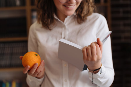 Smiling Teenage girl in glasses holding an opened book with an orange in the other handの写真素材