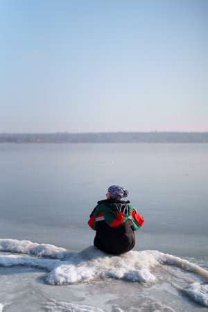 A woman in a colorful thermal costume enjoying winter while sitting and watching a frozen lake with blue sky in the background. Freedomの写真素材