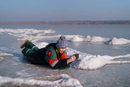 Authentic woman taking pictures with a phone of natural frozen lake with a clear blue sky in background. Explorerの写真素材