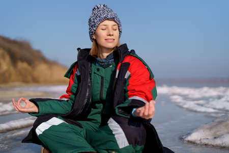 A woman in a colorful thermal costume enjoying winter while sitting in a meditation pose on a frozen lake with blue sky in the background. Freedom, peace, quiet, serenityの写真素材