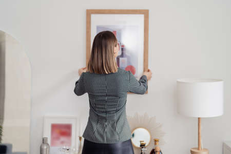Home improvement. Back view of a woman hanging a picture on a white wall in a light interiorの写真素材