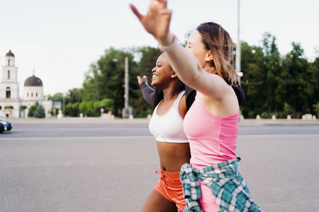 Cheerful smiling friends in sportswear running in the city dicussing. Multiethnic women having a fitness workout.の写真素材