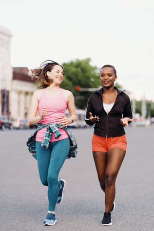 Cheerful smiling friends in sportswear running in the city dicussing. Multiethnic women having a...