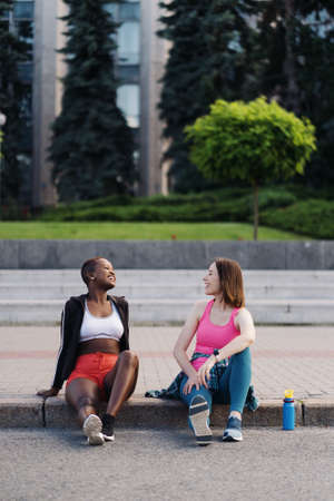 Cheerful smiling friends in sportswear sitting in the city dicussing. Multiethnic women having a fitness workout break.の写真素材