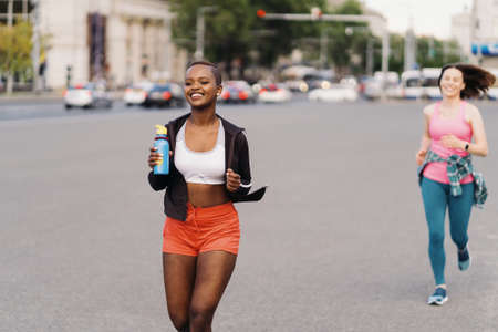 Cheerful smiling friends in sportswear running in the city dicussing. Multiethnic women having a fitness workout.の写真素材