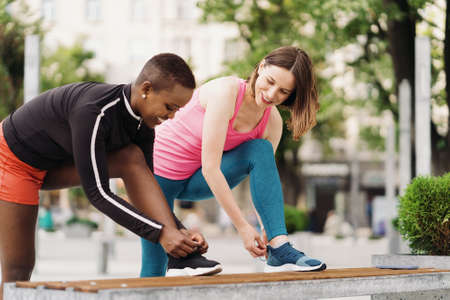 Cheerful smiling friends in sportswear tying sport shoes laces in the city discussing. Multiethnic women having a fitness workout break.の写真素材