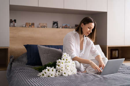 Young woman sitting on the bed wearing pajamas, with pleasure enjoying white flowers, chatting using laptopの写真素材