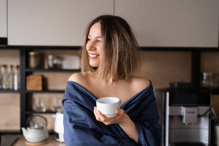 Sleepy positive woman wrapped in blue bed sheet drinking coffee in the morning in the kitchenの写真素材