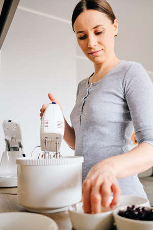 Young woman baking a gem pie in the kitchen standing at the counter in home dress using a handheld mixer to whisk freshest ingredients in a white mixing bowl, ingredients on the tableの写真素材