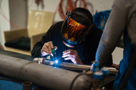 Worker in helmet with flames welding the steel part of a motorbike using an argon welding machine...