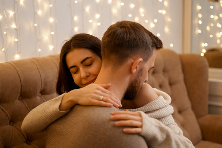 Happy married young couple hugging, sitting on cozy couch together, woman and man enjoying leisure time, relaxing on sofa in living room at home with lights in backgroundの写真素材