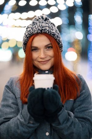 Girl drinking hot coffee while walking in the evening city market decorated with holiday lights in the evening. Feeling happy in big city. Spending winter vacationsの写真素材