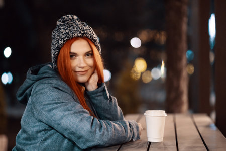 Girl drinking hot coffee while sitting at the table in the evening city market decorated with holiday lights. Feeling happy in big city. Spending winter vacationsの写真素材