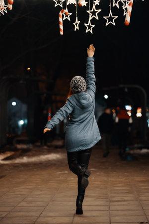 Outdoor night photo of young beautiful happy smiling girl enjoying festive decoration, posing in Christmas fair, in street of european city, wearing knitted beanie hatの写真素材