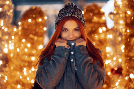 Outdoor night photo of young beautiful happy smiling girl enjoying festive decoration, posing in Christmas fair, in street of european city, wearing knitted beanie hatの写真素材