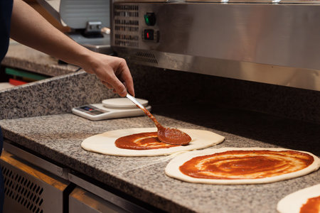 Anonymous male chef spreading tomato sauce with big iron spoon onto pizzaの写真素材