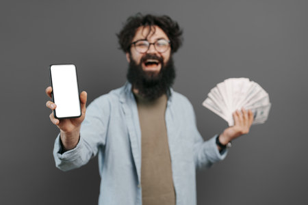 Joyful bearded man holding mobile phone and cash moneyの写真素材