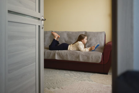 Young girl lying on a sofa engrossed in reading a blue bookの写真素材