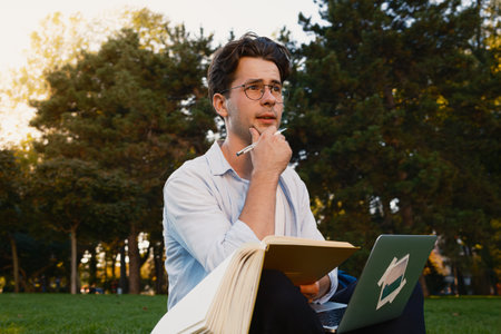 Thoughtful Student Studying Outdoors with Notebook and Laptop in a Park