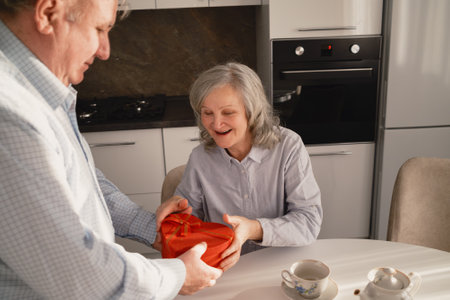 Senior couple exchanging a gift in their cozy kitchen settingの写真素材