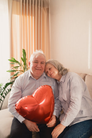 Elderly couple embracing with heart-shaped balloon in warm living room settingの写真素材