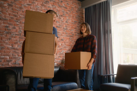 Two people carrying cardboard boxes in a sunny apartment with brick walls.