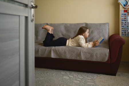 Young girl reading a book while lying on the couch at homeの写真素材