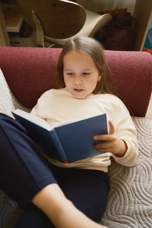 Young girl reading a book comfortably seated indoors on a chairの写真素材