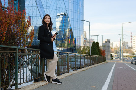 Woman standing outdoors holding a smartphone near modern urban architectureの写真素材