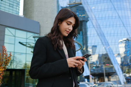 Businesswoman checks her smartphone outside modern office building during work breakの写真素材