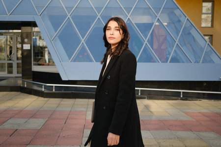 Confident woman posing outdoors in stylish black coat near modern architectureの写真素材