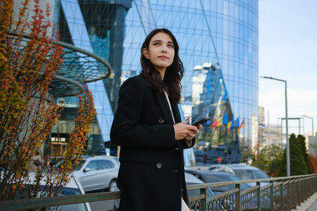 Businesswoman standing outdoors near modern building holding smartphone during daytimeの写真素材