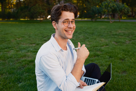 Happy young man working on a laptop and writing in a parkの写真素材