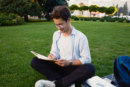 Student sitting on grass writing in a notebook enjoying a sunny day outdoorsの写真素材