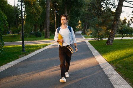 Student walking through a park carrying books during a sunny dayの写真素材