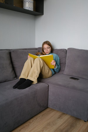 Young girl reading a yellow book while sitting on a gray couchの写真素材