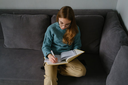 Teenage girl studying on sofa with book and pen in relaxed settingの写真素材