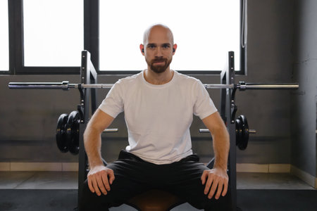Man sitting confidently in gym on bench with weightlifting equipment in backgroundの写真素材