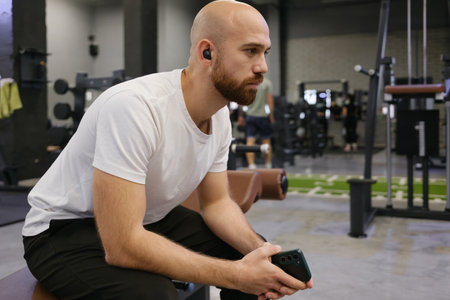 Man with earbuds sitting in a gym while holding a smartphone thoughtfully