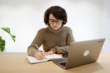 Young woman studying with a notebook and laptop in a bright workspaceの写真素材